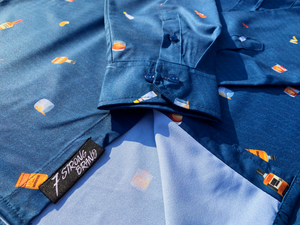 Sweep tag and sleeve view of "Call Me Old Fashioned" adult long sleeve button-down. Features elements of an Old Fashioned cocktail (bottle of bourbon, glasses, oranges, stirrers, and ice) against a blue background which faintly spells out an Old Fashioned recipe. 