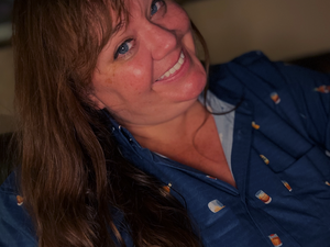 Female model smiling at camera wearing the "Call Me Old Fashioned" adult long sleeve button-down. Features elements of an Old Fashioned cocktail (bottle of bourbon, glasses, oranges, stirrers, and ice) against a blue background which faintly spells out an Old Fashioned recipe. 