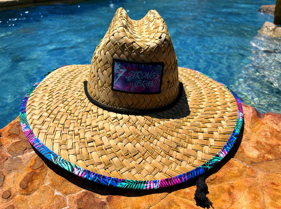 Centered front facing close up of the 7-Strong "Neon Coast" Sun Hat, made from straw, which features the Neon Coast design in a patch on the front, with "7-Strong Brand" written across it. The pattern can also be seen lining the brim of the hat, along with the underside. A black strap keeps the hat in place.