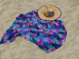 Full centered view of the 7-Strong "Neon Coast" adult button down shirt. Featuring vibrant teal, pink, and blue palm fronds, overlapping throughout the shirt, against a black/deep purple background. The shirt is laid out on the sand of a beach. Alongside the shirt is the 7-Strong "Neon Coast" Sun Hat, made from straw, which features the Neon Coast design in a patch on the front, with "7-Strong Brand" written across it. The pattern can also be seen lining the brim of the hat.