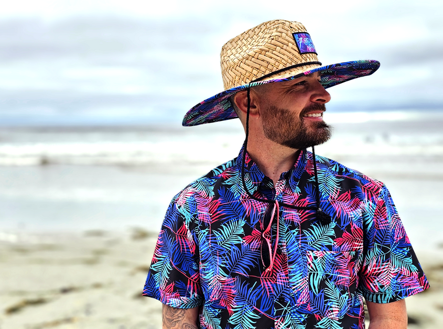 Male model at the beach, smiling and looking off in the distance, wearing the 7-Strong "Neon Coast" Sun Hat, made from straw, which features the Neon Coast design in a patch on the front, with "7-Strong Brand" written across it. The pattern can also be seen lining the brim of the hat, along with the underside. A black strap keeps the hat in place. The hat is in combination with the 7-Strong "Neon Coast" shirt with similar feature design. 