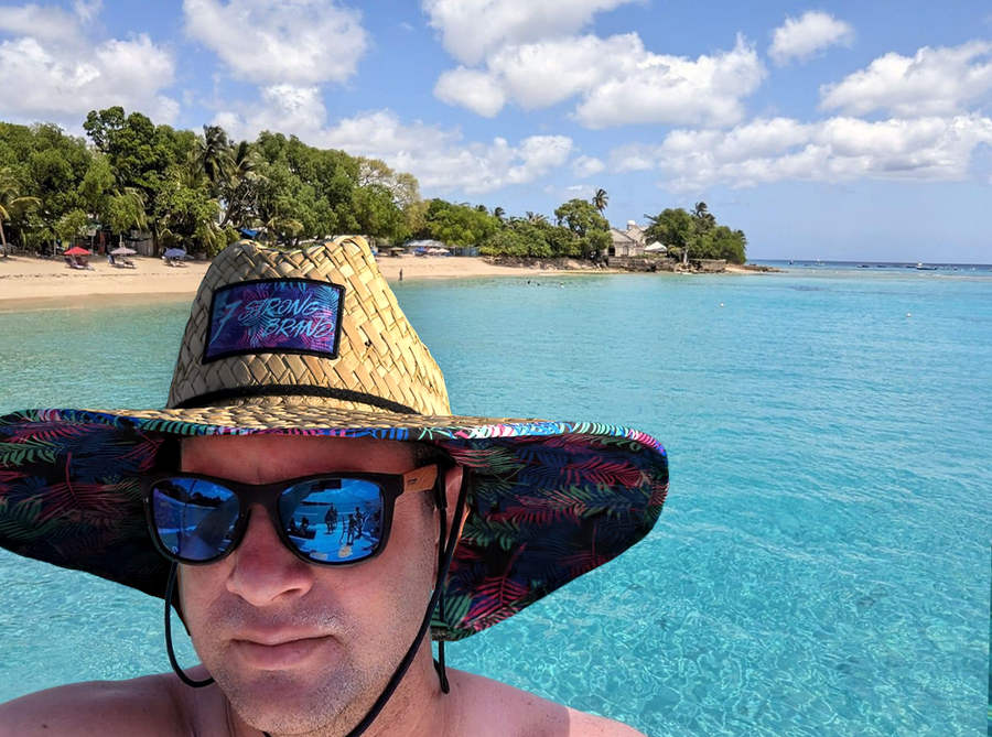 An adult male model in the water, beach coastline behind him, wearing the 7-Strong "Neon Coast" Sun Hat, made from straw, which features the Neon Coast design in a patch on the front, with "7-Strong Brand" written across it. The pattern can also be seen lining the brim of the hat, along with the underside. A black strap keeps the hat in place.