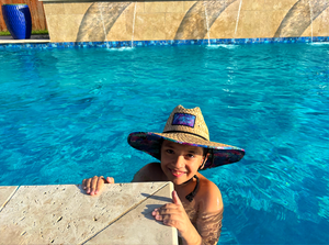 Shot of a child model in a pool wearing the 7-Strong "Neon Coast" Sun Hat, made from straw, which features the Neon Coast design in a patch on the front, with "7-Strong Brand" written across it. The pattern can also be seen lining the brim of the hat, along with the underside. A black strap keeps the hat in place. 