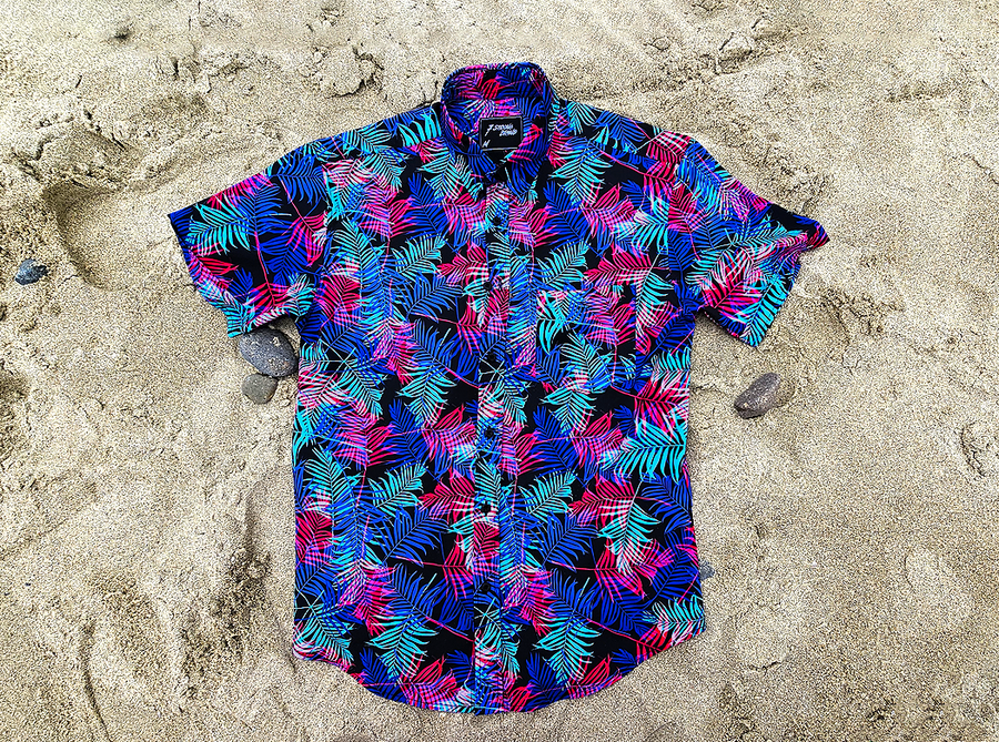 Full centered view of the 7-Strong "Neon Coast" adult button down shirt. Featuring vibrant teal, pink, and blue palm fronds, overlapping throughout the shirt, against a black/deep purple background. The shirt is laid out on the sand of a beach.