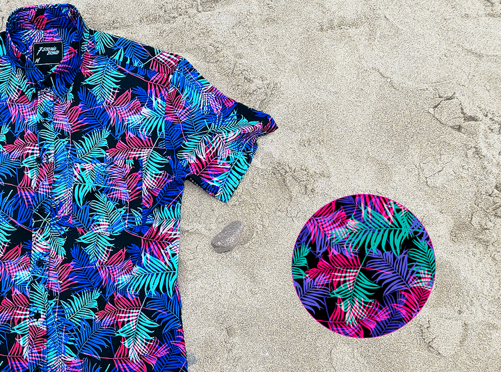 3/4 close up view of the 7-Strong "Neon Coast" adult button down shirt. Featuring vibrant teal, pink, and blue palm fronds, overlapping throughout the shirt,  against a black/deep purple background. The shirt is laid out on the sand of a beach. In the bottom right corner, there is a detail circle featuring a close up of the shirt's design.  