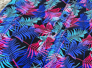 Close-up midsection view of the 7-Strong "Neon Coast" adult button down shirt. Featuring vibrant teal, pink, and blue palm fronds, overlapping throughout the shirt, against a black/deep purple background. The shirt is laid out on the sand of a beach.