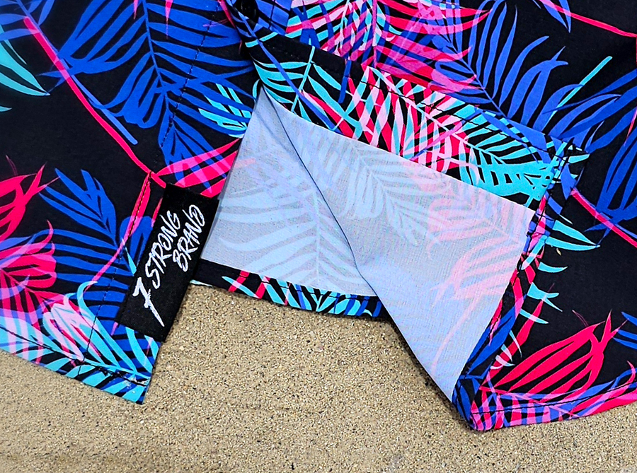 Bottom sweep tag view of the 7-Strong "Neon Coast" adult button down shirt. Featuring vibrant teal, pink, and blue palm fronds, overlapping throughout the shirt, against a black/deep purple background. The shirt is laid out on the sand of a beach.