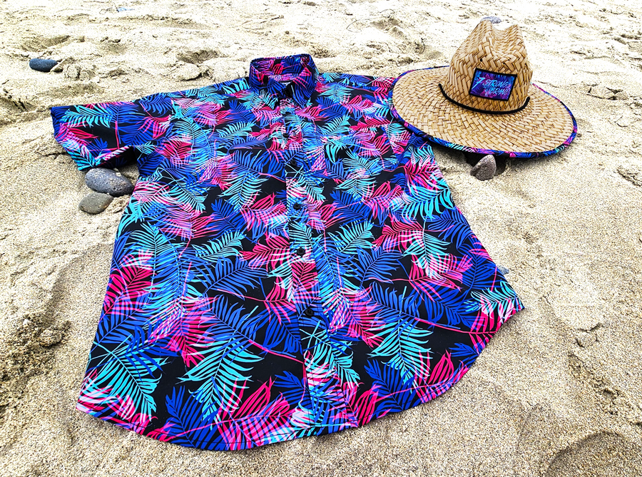 Full centered view of the 7-Strong "Neon Coast" adult button down shirt. Featuring vibrant teal, pink, and blue palm fronds, overlapping throughout the shirt, against a black/deep purple background. The shirt is laid out on the sand of a beach. Alongside the shirt is the 7-Strong "Neon Coast" Sun Hat, made from straw, which features the Neon Coast design in a patch on the front, with "7-Strong Brand" written across it. The pattern can also be seen lining the brim of the hat. 