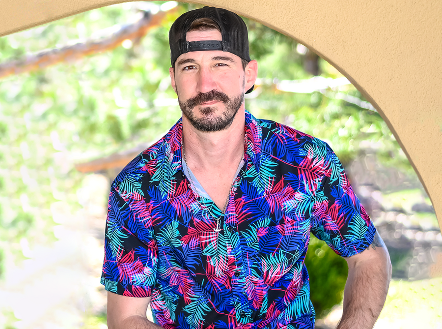 Male model, facing camera with a slight smile, wearing the 7-Strong "Neon Coast" adult button down shirt. Featuring vibrant teal, pink, and blue palm fronds, overlapping throughout the shirt, against a black/deep purple background. Model is seated in an outside setting. 