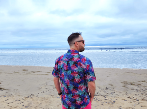 Male model, back to camera looking off in the distance on the beach, wearing the 7-Strong "Neon Coast" adult button down shirt. Featuring vibrant teal, pink, and blue palm fronds, overlapping throughout the shirt, against a black/deep purple background.