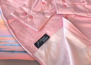 Bottom sweep view of the pink long sleeve EveryWEAR adult button up. Pattern is subtle white 7-Bolts lined up all against each other across a pink background. The shirt is featured on a soft background of a wave hitting the beach. 