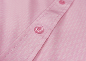 Mid pocket tag view of the pink short sleeve EveryWEAR adult button up. Pattern is subtle white 7-Bolts lined up all against each other across a pink background. The shirt is featured on a soft background of a wave hitting the beach.
