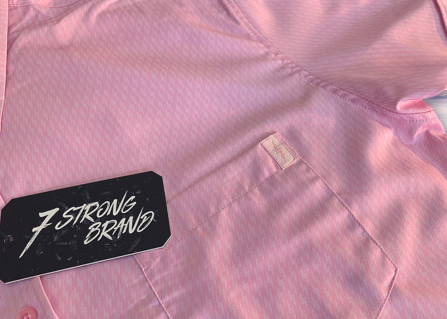 Corner pocket view of the pink short sleeve EveryWEAR adult button up. Pattern is subtle white 7-Bolts lined up all against each other across a pink background. The shirt is featured on a soft background of a wave hitting the beach.