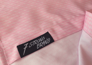 Bottom sweep tag view of the pink short sleeve EveryWEAR adult button up. Pattern is subtle white 7-Bolts lined up all against each other across a pink background. The shirt is featured on a soft background of a wave hitting the beach. 