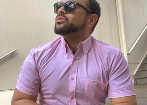 Medium shot of a male model gazing off wearing the pink short sleeve EveryWEAR adult button up. Pattern is subtle white 7-Bolts lined up all against each other across a pink background.