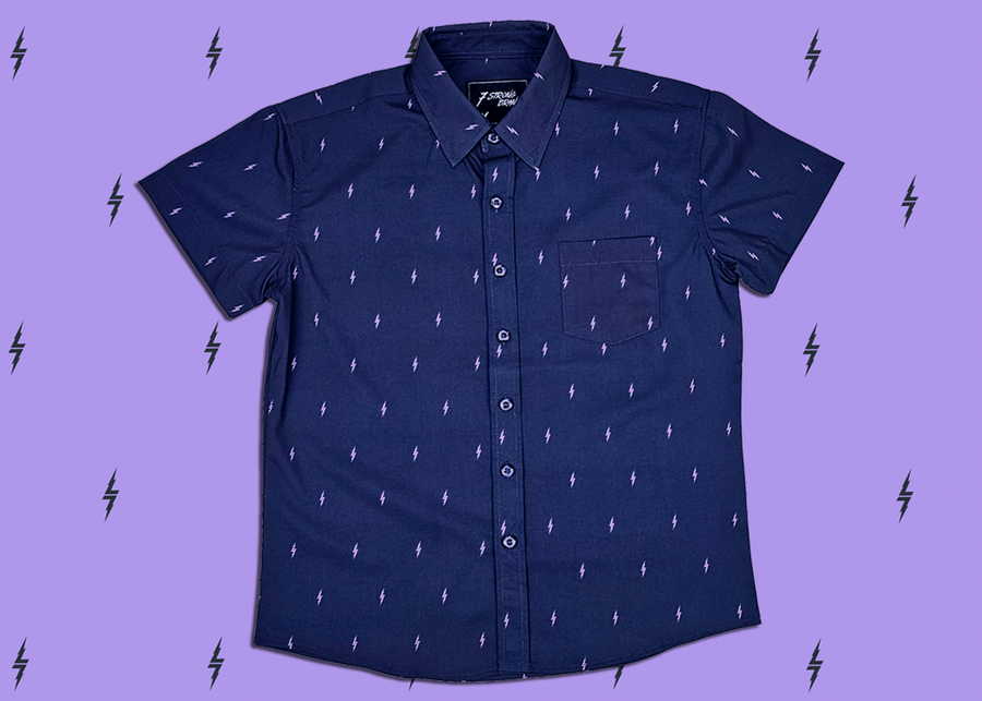 Centered, full view of the youth 7-Strong Slate 7-Bolt short sleeve shirt, featuring lavender 7-bolts interspersed on a slate gray background. The shirt is featured on a lavender backdrop with black bolts.