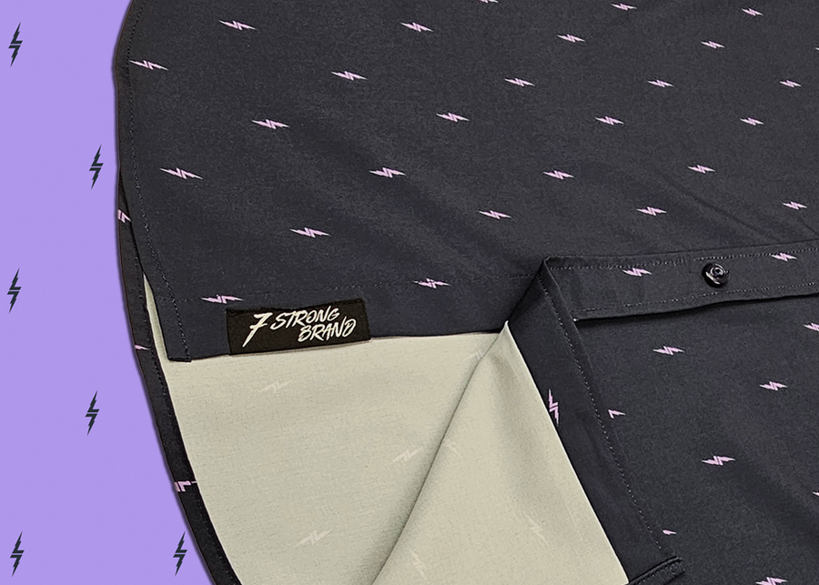 A side angled, close up sweep tag view of the 7-Strong Slate 7-Bolt short sleeve shirt, featuring lavender 7-bolts interspersed on a slate gray background. The shirt is featured on a lavender backdrop with black bolts