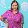 Load image into Gallery viewer, A shot of a female model wearing the 7-Strong adult &quot;Strongfetti&quot; button down, a bold pink shirt featuring black, yellow, lime green, and navy blue scratches that appear like confetti falling from the sky. The model is standing against a colorful bubble design wall peering into the camera.
