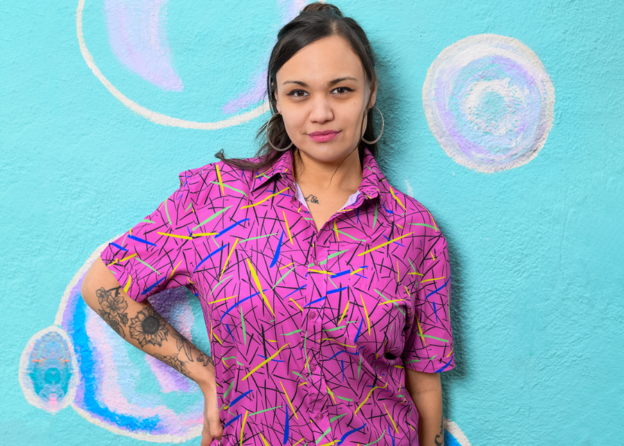 A shot of a female model wearing the 7-Strong adult "Strongfetti" button down, a bold pink shirt featuring black, yellow, lime green, and navy blue scratches that appear like confetti falling from the sky. The model is standing against a colorful bubble design wall peering into the camera.
