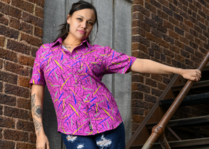 A shot of a female adult model wearing the 7-Strong adult "Strongfetti" button down, a bold pink shirt featuring black, yellow, lime green, and navy blue scratches that appear like confetti falling from the sky. The model is standing on stairs, leaning against a brick wall looking into camera. 