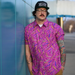 Load image into Gallery viewer, A shot of a male adult model wearing the 7-Strong adult &quot;Strongfetti&quot; button down, a bold pink shirt featuring black, yellow, lime green, and navy blue scratches that appear like confetti falling from the sky. The model is standing against a blue exterior wall peering past the camera.
