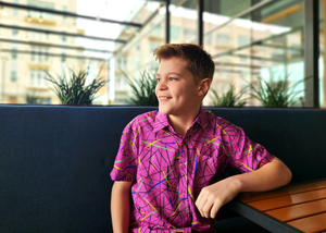 A shot of a male youth model wearing the 7-Strong youth "Strongfetti" button down, a bold pink shirt featuring black, yellow, lime green, and navy blue scratches that appear like confetti falling from the sky. The model is sitting at table smiling and looking off. 