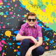 Load image into Gallery viewer, A shot of a male youth model crouched and smiling towards camera wearing the 7-Strong youth &quot;Strongfetti&quot; button down, a bold pink shirt featuring black, yellow, lime green, and navy blue scratches that appear like confetti falling from the sky.  Model is displayed against a multi-colored paint splatted wall design. 
