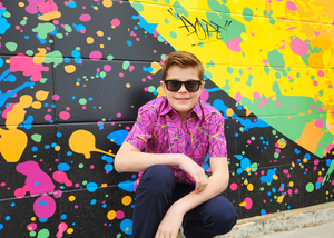 A shot of a male youth model crouched and smiling towards camera wearing the 7-Strong youth "Strongfetti" button down, a bold pink shirt featuring black, yellow, lime green, and navy blue scratches that appear like confetti falling from the sky.  Model is displayed against a multi-colored paint splatted wall design. 