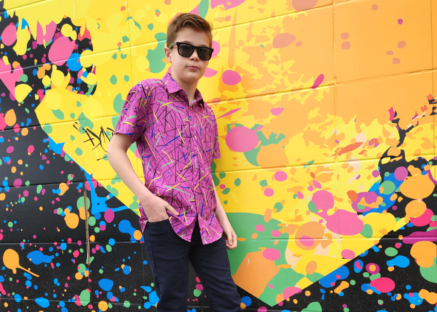 A shot of a male youth model wearing the 7-Strong youth "Strongfetti" button down, a bold pink shirt featuring black, yellow, lime green, and navy blue scratches that appear like confetti falling from the sky. The model is standing against a colorful paint-splatter design wall peering into the camera. 