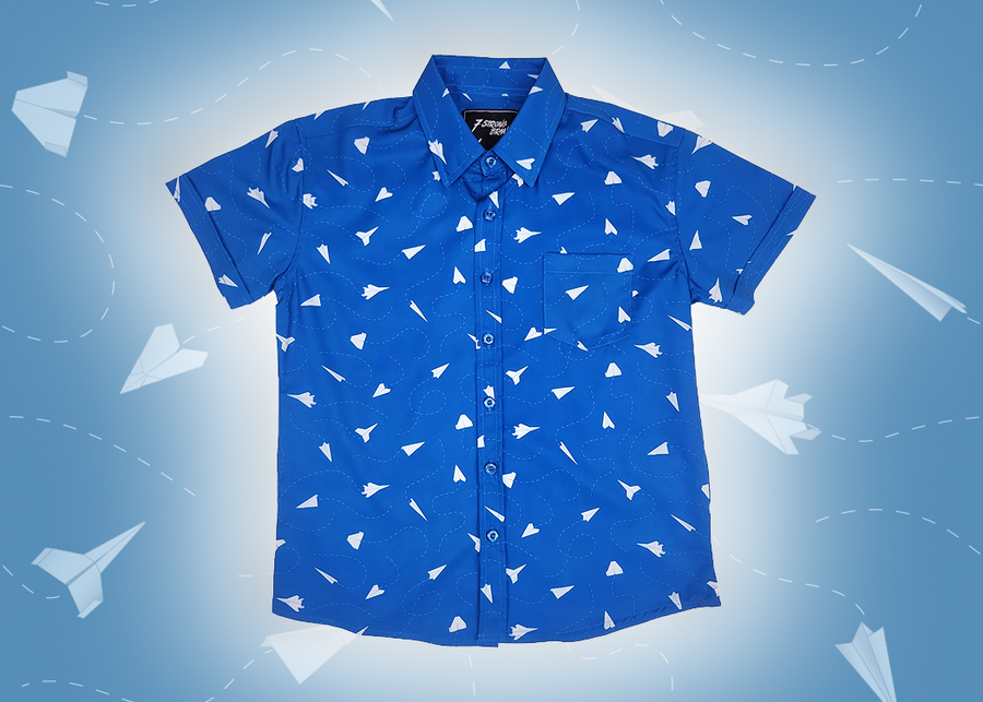 A full view of the youth 7-Strong "Paper Planes" button down shirt - featuring various sizes and designs of paper airplanes, some with dotted trails behind them against a royal blue background. The shirt is displayed against a faced background of the shirt details.
