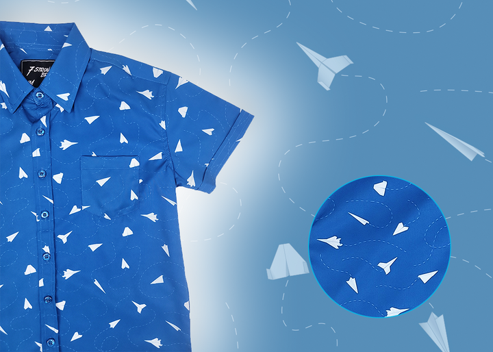 A 3/4 view of the youth 7-Strong "Paper Planes" button down shirt - featuring various sizes and designs of paper airplanes, some with dotted trails behind them against a royal blue background. The shirt is displayed against a faced background of the shirt details. In the bottom right is a detail circle with a close up of the design. 