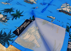 Bottom sweep tag view of the 7-Strong "7-Seas" shirt, a bright blue colored shirt with various nautical depictions such as islands, ships, mermaids, etc - drawn in a treasure map like fashion. The shirt is displayed against a weathered treasure map and compass background.