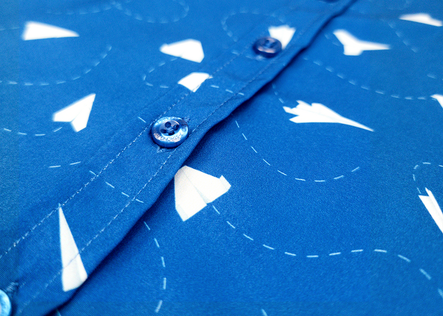 A mid-button view of the youth 7-Strong "Paper Planes" button down shirt - featuring various sizes and designs of paper airplanes, some with dotted trails behind them against a royal blue background. The shirt is displayed against a faced background of the shirt details.