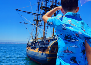A medium close up on a youth model wearing the 7-Strong "7-Seas" shirt, a bright blue colored shirt with various nautical depictions such as islands, ships, mermaids, etc - drawn in a treasure map like fashion. Model is facing the water where a ship is docked, model is looking out towards it. 