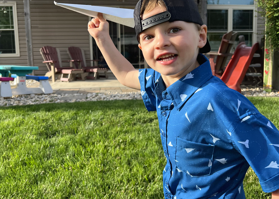A male child model wearing the youth 7-Strong "Paper Planes" button down shirt - featuring various sizes and designs of paper airplanes, some with dotted trails behind them against a royal blue background. The model is holding a paper airplane, smiling at camera, ready to throw the paper airplane. 