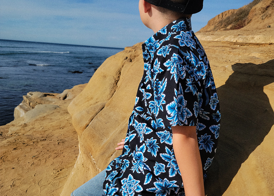 Side view of youth model sitting on rocks wearing the 7-Strong youth Electric Palms shirt, a collection of electric blue palm fronds featured against a black shirt. Model is overlooking the water. 