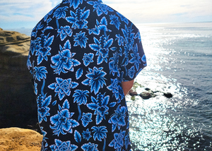 Back view of a youth model wearing the 7-Strong youth Electric Palms shirt, a collection of electric blue palm fronds featured against a black shirt. Model is displayed against an overlook of a scenic beach.
