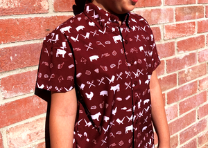 Medium close up of a youth male model against a brick wall wearing the 7-Strong youth "Smoke Show" button up, a deep maroon red colored shirt with rows of various barbecue and cookout related items and delicacies silhouetted in white.
