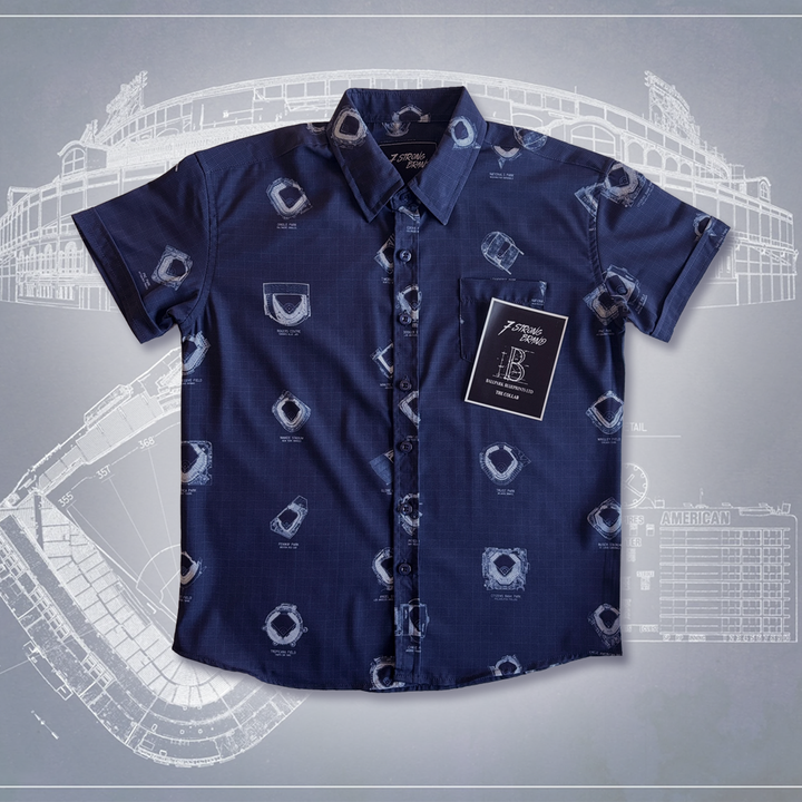 7-Strong's Ballpark Blueprints collaboration featuring a navy blue button-down shirt with an all-over print featuring white blueprint-style diagrams of various Major League Baseball stadiums. The shirt is laid flat on a background that also displays similar stadium blueprints. 