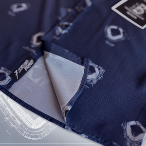 A close-up shot of the shirt's fabric, showing the detailed ballpark blueprints and a folded corner revealing a lighter-colored lining. A "7 Strong Brand" label is visible on the inner seam.