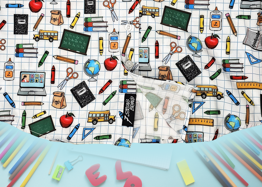 Bottom sweep tag view of 7-Strong Class Act  youth button-down featuring various classroom items such as pencils, rulers, globes, notebooks, apples, protractors, etc against a white graph paper background. Shirt is against a background of school supplies. 