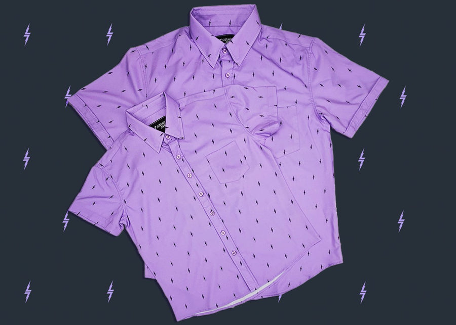 Full centered view of the adult and youth 7-Strong Deep Lavender 7-Bolt short sleeve shirts overlapping each other, featuring black 7-bolts interspersed on a deep lavender background. The shirt is featured on a slate backdrop with lavender bolts. 