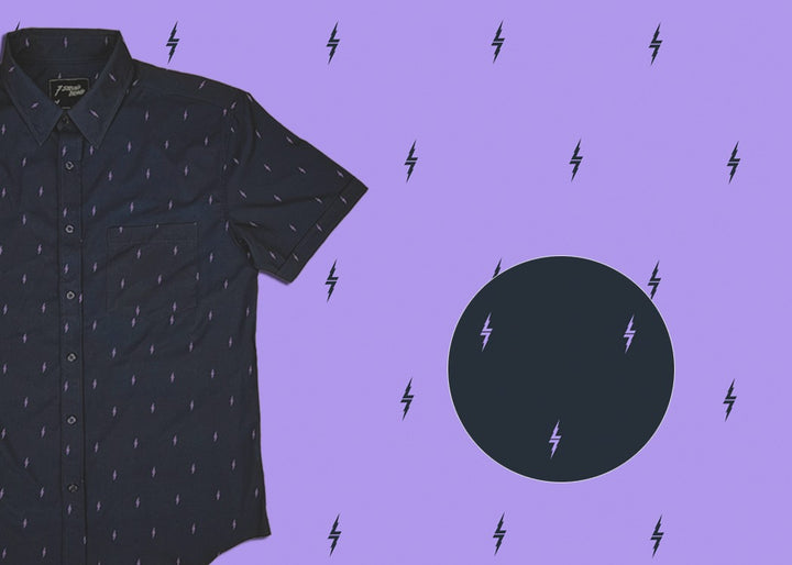 3/4 view of the 7-Strong Slate 7-Bolt short sleeve shirt, featuring lavender 7-bolts interspersed on a slate gray background. The shirt is featured on a lavender backdrop with black bolts. The bottom left features a detail circle showing the 7-bolt design up close.