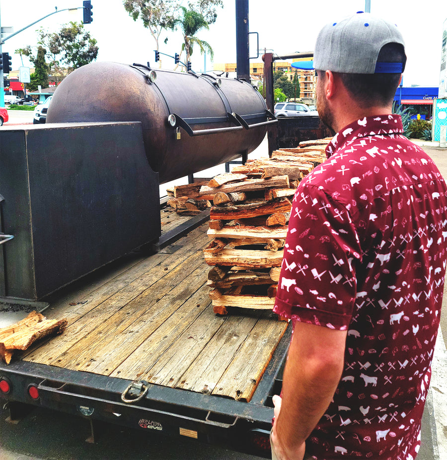 A male model, from the back, looking at a BBQ smoker wearing the 7-Strong adult "Smoke Show" button up, a deep maroon red colored shirt with rows of various barbecue and cookout related items and delicacies silhouetted in white.