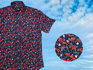 3/4 full view of the 7-Strong "Aye Poppy" button down, featuring an array of red poppys with white sprigs on a deep navy blue shirt. The shirt is displayed against a partly cloudy sky. The bottom right has a detail circle featuring a close up of the design. 