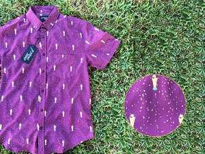 3/4 Full view of the Cup of Life adult button up, a off-maroon colored shirt decorated throughout with falling gold confetti and depictions of the World Cup statue. The shirt is laid out on a grass pitch. In the bottom right corner is a detail circle, highlighting the shirt's design. 