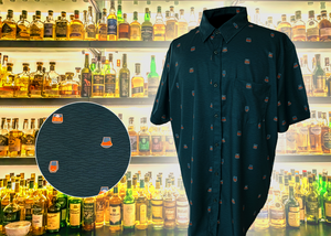 Full display view of the 7-Strong "Straight Sippin'" Short Sleeve button down. The shirt is a deep green with black ripples as its background, while the foreground has various drinking glasses with various levels of whiskey, some with ice, others without. The shirt is shown against a background of various whiskies and bourbons on a shelf. In the left corner, there is a detail circle showcasing the shirt's glass designs, up close. 