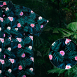 Load image into Gallery viewer, 3/4 view of the Adult 7-Strong &quot;Tropic Like It&#39;s Hot&quot; button down shirt - a black shirt with distinctive green palm trees and pink, white, and purple flowers peering out from behind the palms. Shirt is shown on a tropical rainforest background. Bottom right corner features a circle that details the flowers and palms designed on the shirt in close up. 

