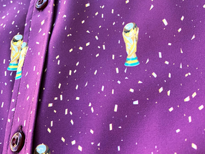 Middle button close up view of the Cup of Life adult button up, a off-maroon colored shirt decorated throughout with falling gold confetti and depictions of the World Cup statue. The shirt is laid out on a grass pitch. 