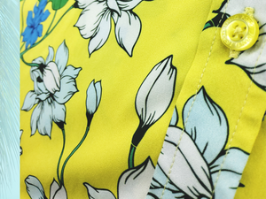 Close up mid drift button view of the 7-Strong "Late Bloomer" adult button down - a yellow background shirt with white and blue flowers patterned all over it. Shirt is against a blue tropical sky with palm trees in the distance. 